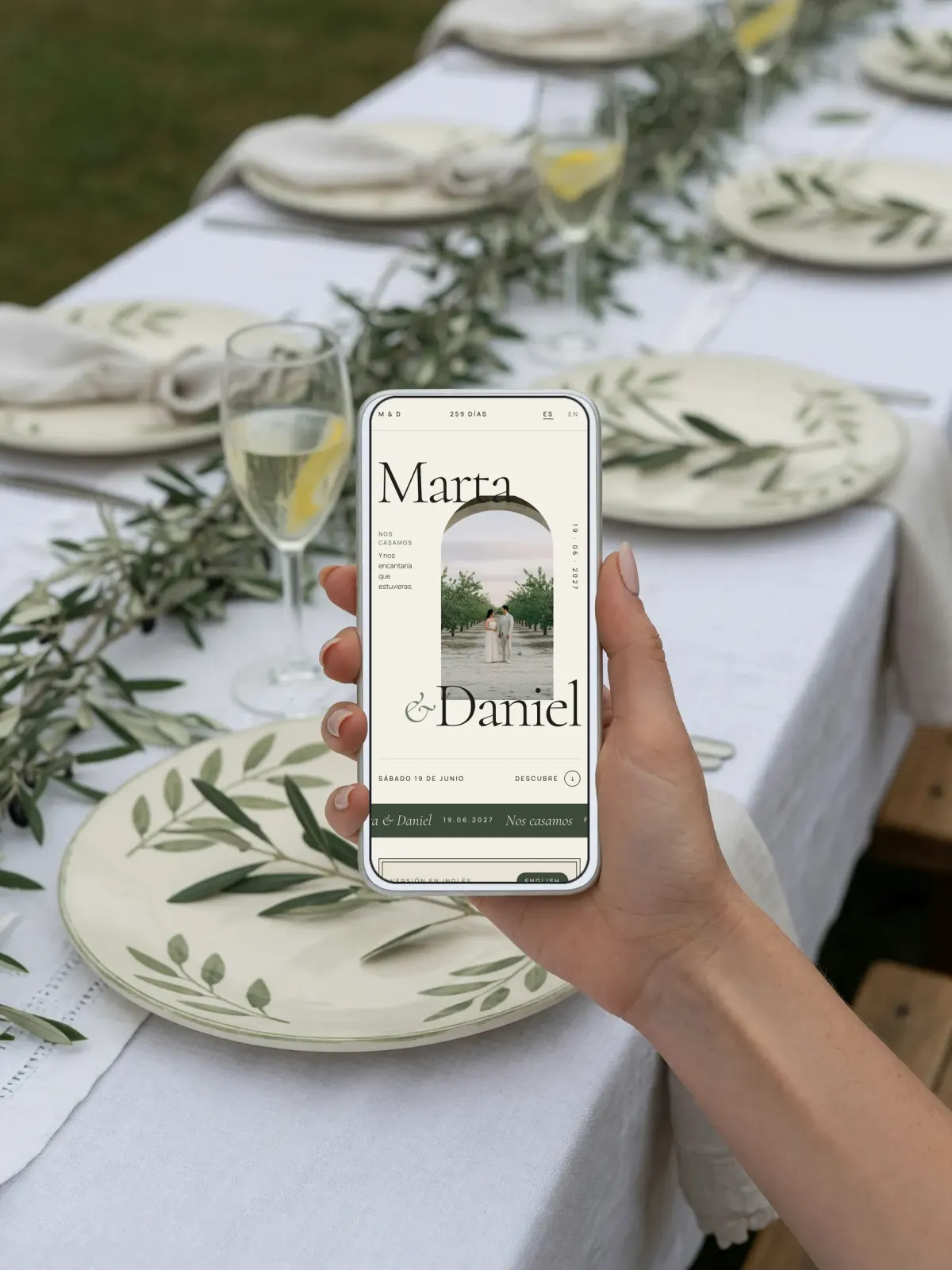 Un invitado sostiene el móvil sobre la mesa del banquete, entre platos pintados con ramas de olivo y copas de vino blanco